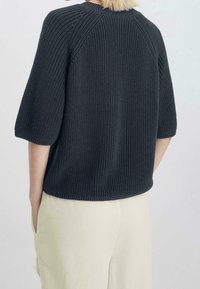 Personne aux cheveux blonds courts portant un pull à manches courtes en tricot gris foncé et un pantalon beige clair, mains dans les poches, vue de dos.