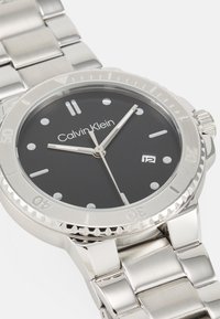 Reloj de acero inoxidable con una esfera negra, manecillas plateadas y marcadores de hora. Presenta una pantalla de fecha y un bisel giratorio. Nombre de la marca mostrado de forma prominente.
