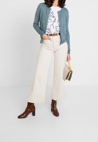 Groene cardigan over een wit T-shirt met print, beige broek met wijde pijpen en bruine enkellaarsjes met hak. Draagt een kleine beige handtas met accenten.