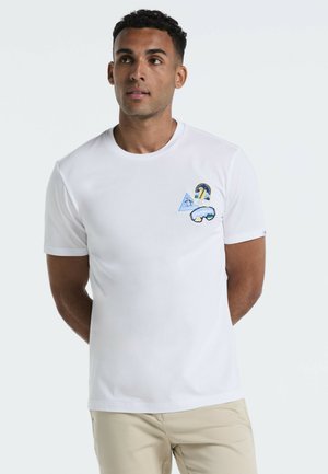 Mann steht mit hinter dem Rücken verschränkten Händen, trägt ein weißes T-Shirt mit Pinguin-Patches und beige Hosen vor einem weißen Hintergrund.