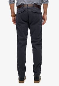 Uomo che indossa pantaloni blu navy, cintura di pelle marrone, camicia a quadri blu e scarpe nere, in piedi di spalle su sfondo bianco.