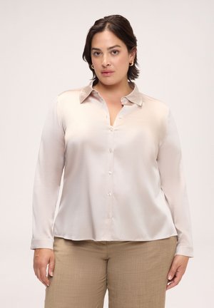 Frau mit kurzen dunklen Haaren trägt eine hellbeige satinierte Knopfleiste-Bluse und hellbraune Hose vor einem schlichten hellen Hintergrund.