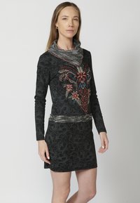 Schwarz langärmliges Kleid mit strukturiertem Blumenmuster, das bunte Stickereien auf der Brust sowie einen mit Metall akzentuierten Ausschnitt und Taillenbereich aufweist.