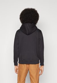 Fekete kapucnis pulóver, bordázott mandzsettákkal és hem-vel, egyszínű textúrával. Barna nadrággal viselve, hátulról nézve.