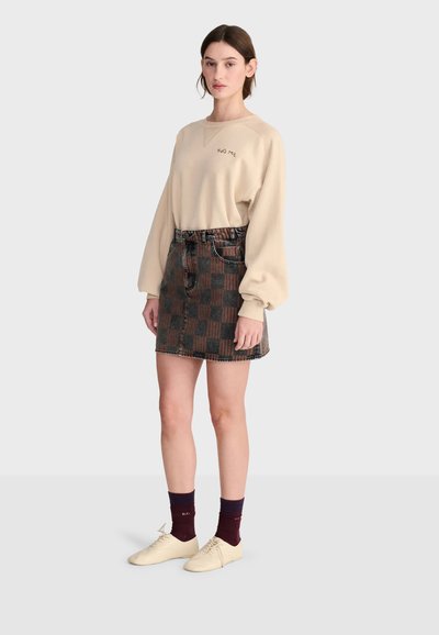 Jeune femme portant un sweat beige "Hug Me", une jupe à carreaux marron et noire, des chaussettes foncées et des chaussures beiges, debout devant un fond uni.