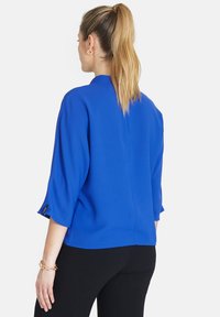 Ljusblå blus med trekvartslånga ärmar och rund halsringning, med en mjuk textur och en liten svart etikettdetalj på ärmen.