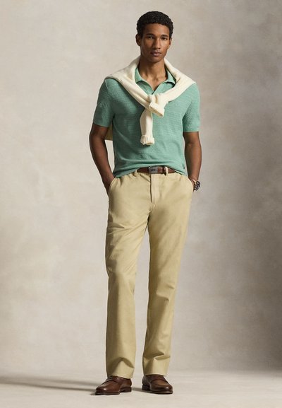 Mężczyzna ubrany w jasnozieloną koszulkę polo z krótkim rękawem, beżowe spodnie, brązowy pasek i buty, z białym swetrem zarzuconym na ramiona, stoi z rękami w kieszeniach.