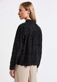 Blusa nera in broderie anglaise con motivi floreali, orlo scallop e maniche lunghe, realizzata in tessuto leggero con una vestibilità rilassata.