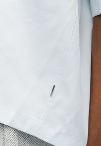 Λευκό ύφασμα ρούχο με πλαϊνό ραφή και μικρή ορθογώνια μεταλλική ετικέτα κοντά στο στρίφωμα, φορεμένο πάνω από υφασμάτινο ύφασμα με γκρι και λευκό ανάγλυφο σχέδιο.