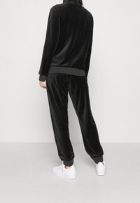 Osoba oblečená v černém velurovém teplákovém soupravě s hřebíkovanými manžetami a bílými teniskami, stojící na neutrálním světle šedém pozadí.