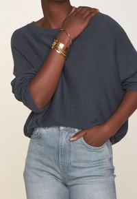Granatowy dziergany sweter z szerokim dekoltem, zestawiony z jasnoniebieskimi, wysokim stanem jeansami. Na jednym nadgarstku złote bransoletki, które dodają metalicznego akcentu.