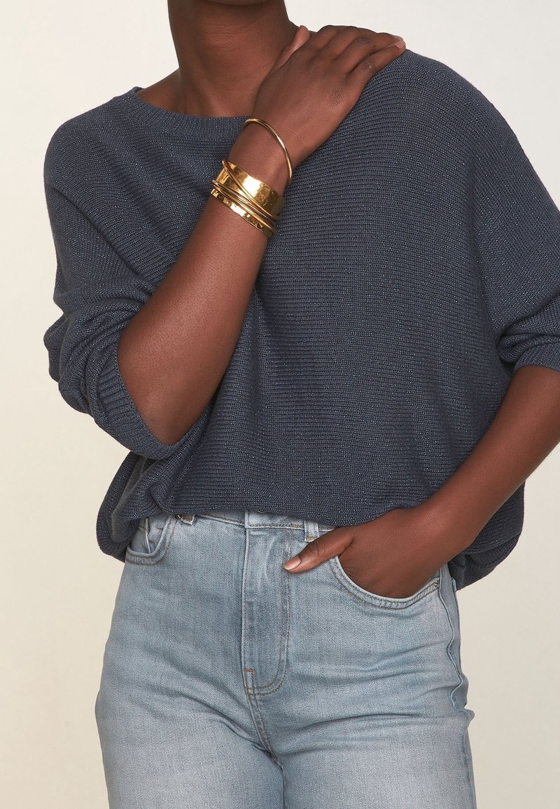 Granatowy dziergany sweter z szerokim dekoltem, zestawiony z jasnoniebieskimi, wysokim stanem jeansami. Na jednym nadgarstku złote bransoletki, które dodają metalicznego akcentu.