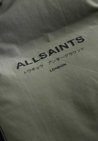 Alyvmedžio spalvos audinys su lygiu paviršiumi, kuriame bold juodu spaudiniu išspausdintas tekstas "ALLSAINTS" ir "LONDON". Po juo yra japoniški simboliai.