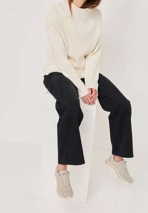 Person iført cremeknittrøje, sorte bukser med vide ben og beige sneakers, siddende på en hvid rektangulær blok mod en ensfarvet baggrund.