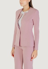 Donna che indossa un blazer aderente di un rosa polveroso e pantaloni abbinati su una maglietta bianca decorata con perline a forma di perla, in piedi su uno sfondo neutro.