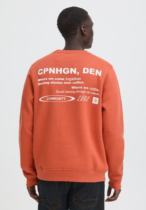 Hombre de espaldas que lleva una sudadera naranja con texto blanco sobre la comunidad y Copenhague en la espalda, sobre un fondo claro y liso.
