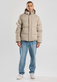 Giacca puffer beige con cappuccio, caratterizzata da un design trapuntato e chiusura a pressione. Indossata sopra jeans azzurri chiari e scarpe da ginnastica bianche.