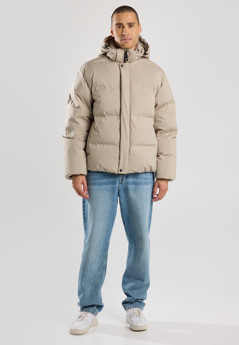 Giacca puffer beige con cappuccio, caratterizzata da un design trapuntato e chiusura a pressione. Indossata sopra jeans azzurri chiari e scarpe da ginnastica bianche.