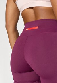 Augstās jostasvietas purpura legingi, izgatavoti no elastīga materiāla, ar sarkanu Nike logotipu uz jostas. Gluda tekstūra un pieguļošs dizains.