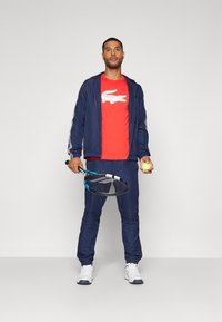 Marine sportsjakke med hvite detaljer, rød t-skjorte med hvit logo, marinebukser og hvite joggesko. Holder en tennisracket og ball.