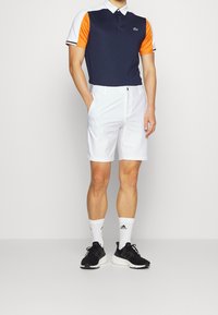 Herrpolo med marinblå framsida, orange ärmar och vita detaljer, i kombination med vita shorts. Accessoarer inkluderar svarta träningsskor och vita strumpor.