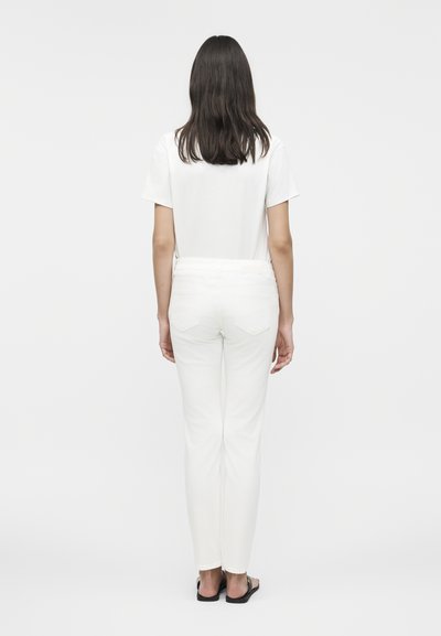 Femme debout de dos portant une chemise blanche à manches courtes, un pantalon blanc et des sandales noires, sur un fond clair uni.