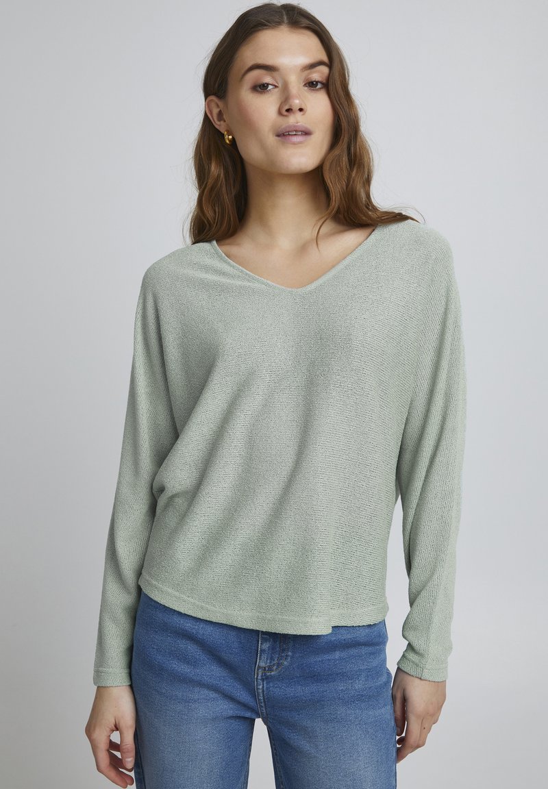 Helles grünes, langärmliges V-Ausschnitt-Pullover aus weichem, strukturiertem Stoff. Getragen mit blauen Jeans, die einen lockeren Schnitt und einen leicht verkürzten Saum aufweisen.