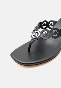 Μαύρο δερμάτινο σανδάλι τύπου σαγιονάρα με κυκλικές εγκοπές που φέρουν λογότυπα "MK" και ασημένιο έμβλημα "MK" στο λουράκι κοντά στα δάχτυλα.
