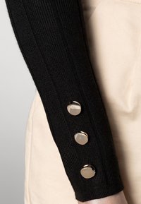 Top negro de manga larga con costillas, con tres botones brillantes de tono plateado en la muñeca, sobre un fondo color crema.