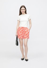 T-shirt branca de mangas curtas combinada com calções vermelhos com padrão floral. Modelo segura uma pequena mala preta, usando sapatos planos pretos.