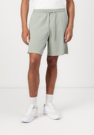 Uomo in piedi su sfondo bianco, indossa pantaloncini grigio chiaro con coulisse, calzini bianchi, sneakers bianche e una maglietta bianca.