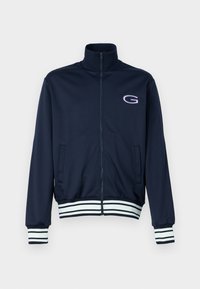Giacca da track in nylon di colore navy con colletto alto, chiusura frontale con zip a tutta lunghezza, tasche laterali e polsini e orlo a righe nei toni bianco e navy. Presenta un logo sul petto.