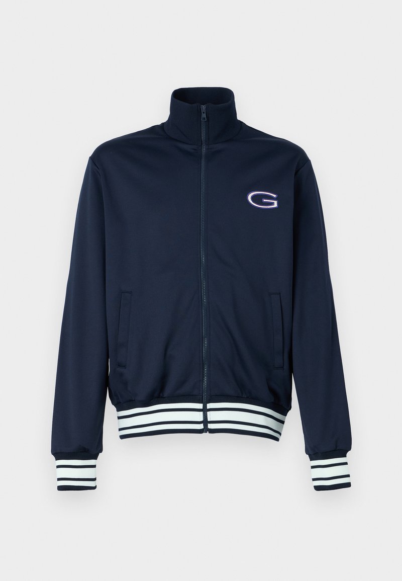 Chaqueta de chándal de marina con cuello alto, cierre frontal de cremallera completa, bolsillos laterales y puños y dobladillo a rayas en blanco y acentos en navy. Presenta un logotipo en el pecho.