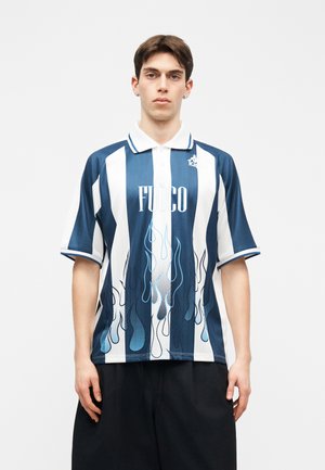 Joven con camiseta polo de rayas verticales azules y blancas con el texto "FUOCO" y gráficos de llamas, combinado con pantalones cortos negros, está de pie quieto.