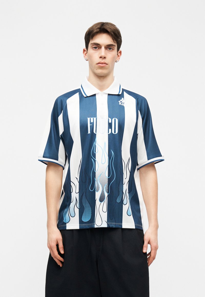 Νέος άνδρας που φοράει μπλε και άσπρη κατακόρυφη ριγωτή πόλο μπλούζα με το κείμενο "FUOCO" και γραφικά φλόγας, συνδυασμένη με μαύρες βερμούδες, στέκεται ακίνητος.