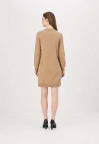 Abito maglione beige a maglia con maniche lunghe, dettaglio a costine ai polsini e all'orlo, spacchi laterali e scollatura rotonda, indossato con tacchi neri.