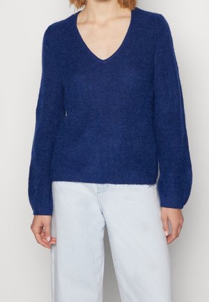Marineblauwe gebreide trui met V-hals, lange mouwen en een textuurfinish. Gekleed met lichte denim jeans voor contrast.