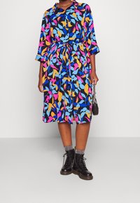 Skjortklänning med svart bakgrund som har färgglada bladmönster i rosa, blått och gult. Inkluderar ett bälte och trekvartslånga ärmar.