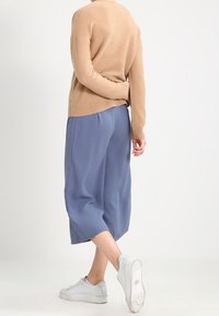 Pullover beige a maglia con texture a coste, abbinato a pantaloni blu larghi e a 3/4, e sneakers grigie con suola testurizzata.