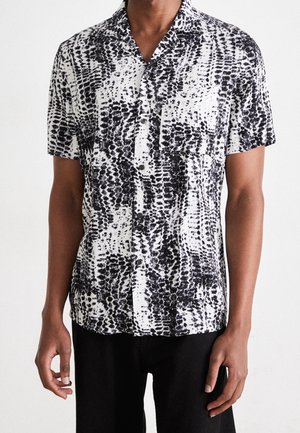 Homme portant une chemise à manches courtes à boutons avec un motif abstrait tacheté noir et blanc et une poche poitrine, associée à un pantalon noir.