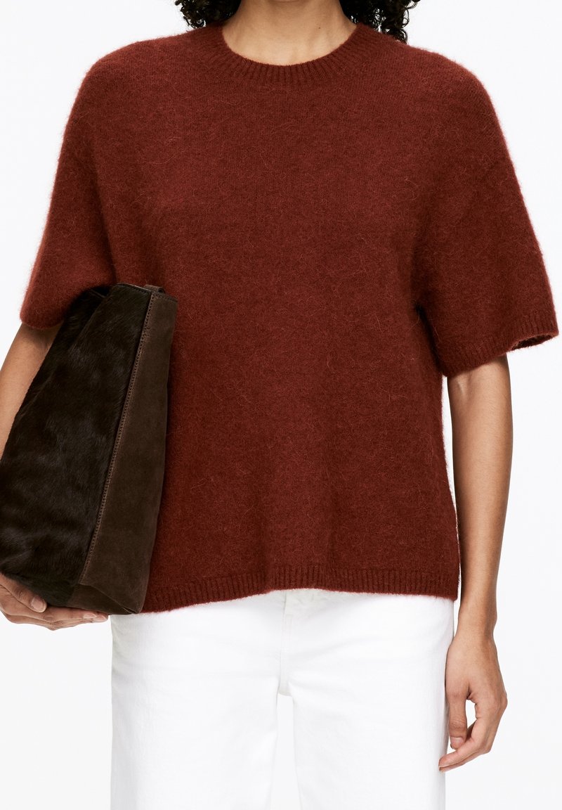 Personne portant un pull à manches courtes duveteux couleur rouille et un pantalon blanc, tenant un grand sac fourre-tout texturé marron foncé.