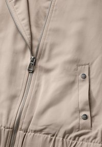 Béžová látka bundy s částečně rozepnutým stříbrným zipem a dvěma stříbrnými druky na vertikálním textilním poutku.