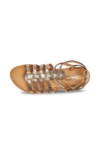 Sandalo strappy metallico oro con suola piatta colorata beige, che presenta un design incrociato e una cinghia alla caviglia per una vestibilità sicura.