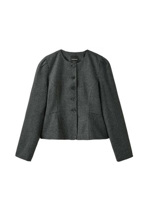 Jeune femme aux longs cheveux bruns se tenant devant un fond gris, portant un blazer court, un pantalon large, tenant un grand sac bleu et des chaussures grises à lacets.