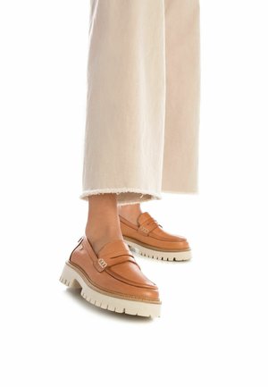 Mocassini in pelle marrone con una spessa suola bianca platform, caratterizzati da due cinturini e dettagli metallici, abbinati a pantaloni chiari con orlo sfrangiato.