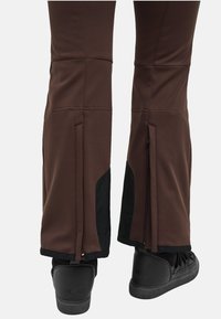 Brune bukser med sorte forstærkede indvendige manchetter og baglommer, båret over sorte lædersneakers med høje såler.