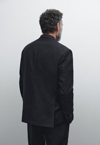 Schwarze Anzugjacke aus maßgeschneidertem Stoff, von hinten betrachtet, mit glattem Material, strukturierten Schultern und einer einzelnen hinteren Belüftung. Ordnungsgemäße Nahtdetails sind sichtbar.