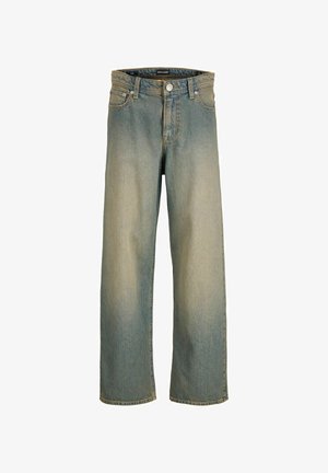 Chłopiec w białej koszulce, wyblakłych niebieskich dżinsach, beżowych butach i granatowej czapce, stoi i patrzy na bok na jednolitym tle.