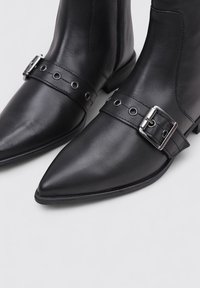 Bottes noires en cuir à bout pointu avec une sangle décorative et une boucle en argent sur le devant, présentées sur un fond clair.