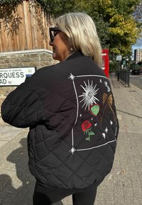 Fekete steppelt dzseki színes hímzett mintákkal, köztük rózsa, nap és absztrakt formák, valamint leopárdmintás díszítéssel.